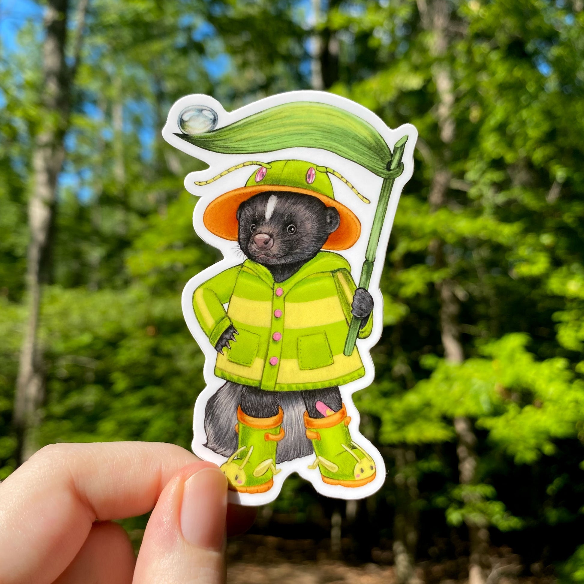 a sticker of a skunk with rainboots and in a green and yellow striped raincoat. He's holding a blade of grass.