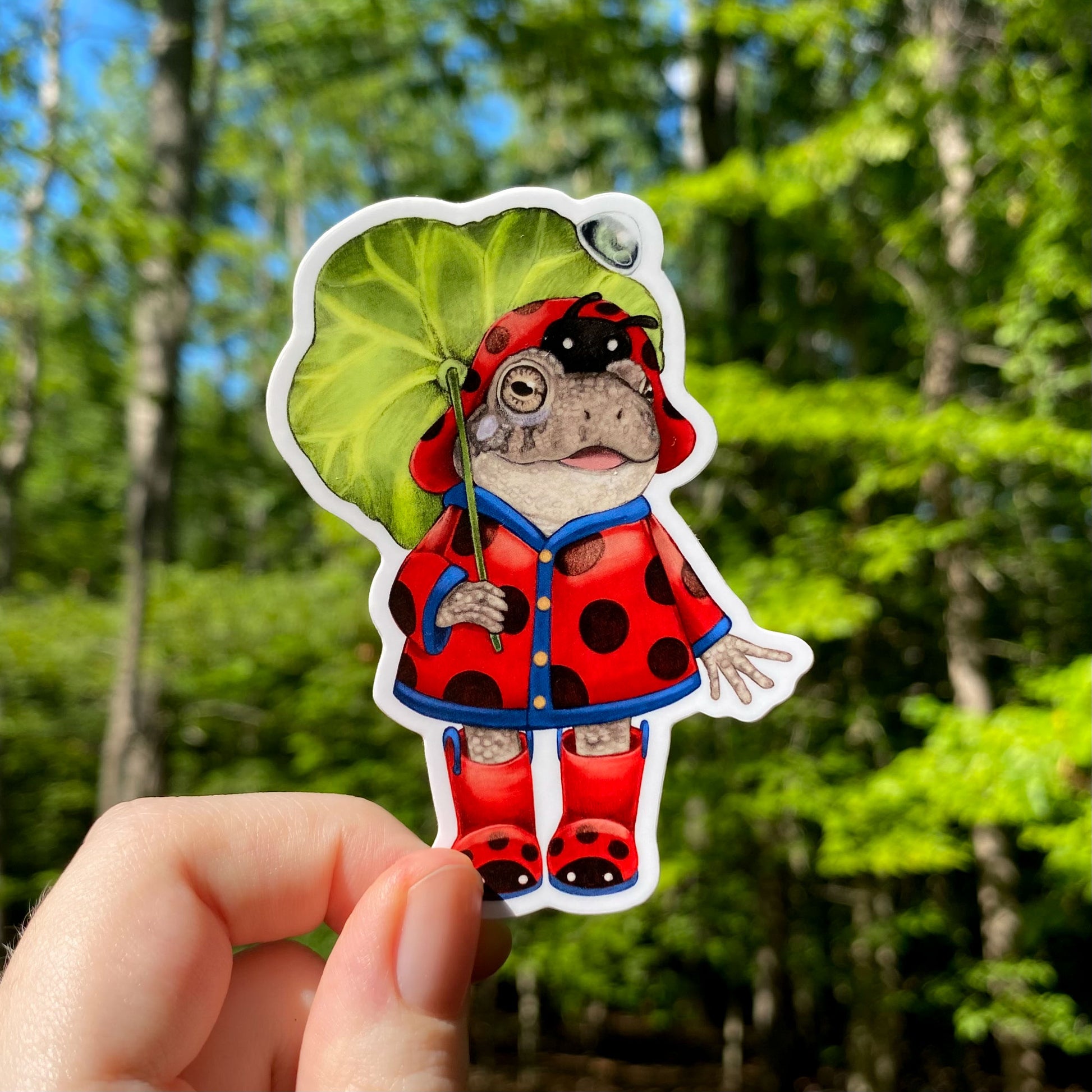 A sticker of a toad in a red ladybug rain coat. He's holding a leaf umbrella.