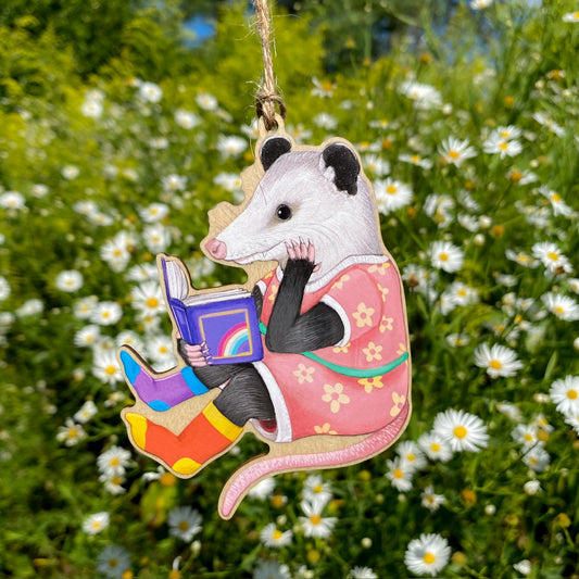 Decorative ornament of an opossum in a pink dress reading a book with a rainbow on a green background.