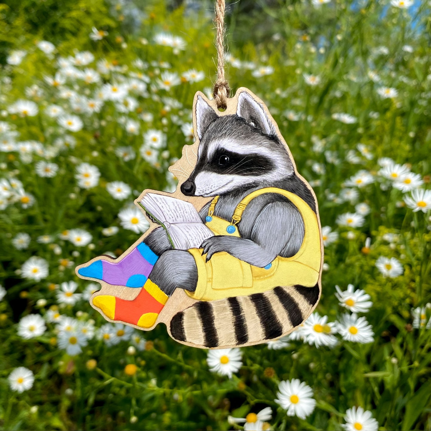 Decorative ornament of a raccoon reading a book with colorful socks, hanging against a natural background with flowers.