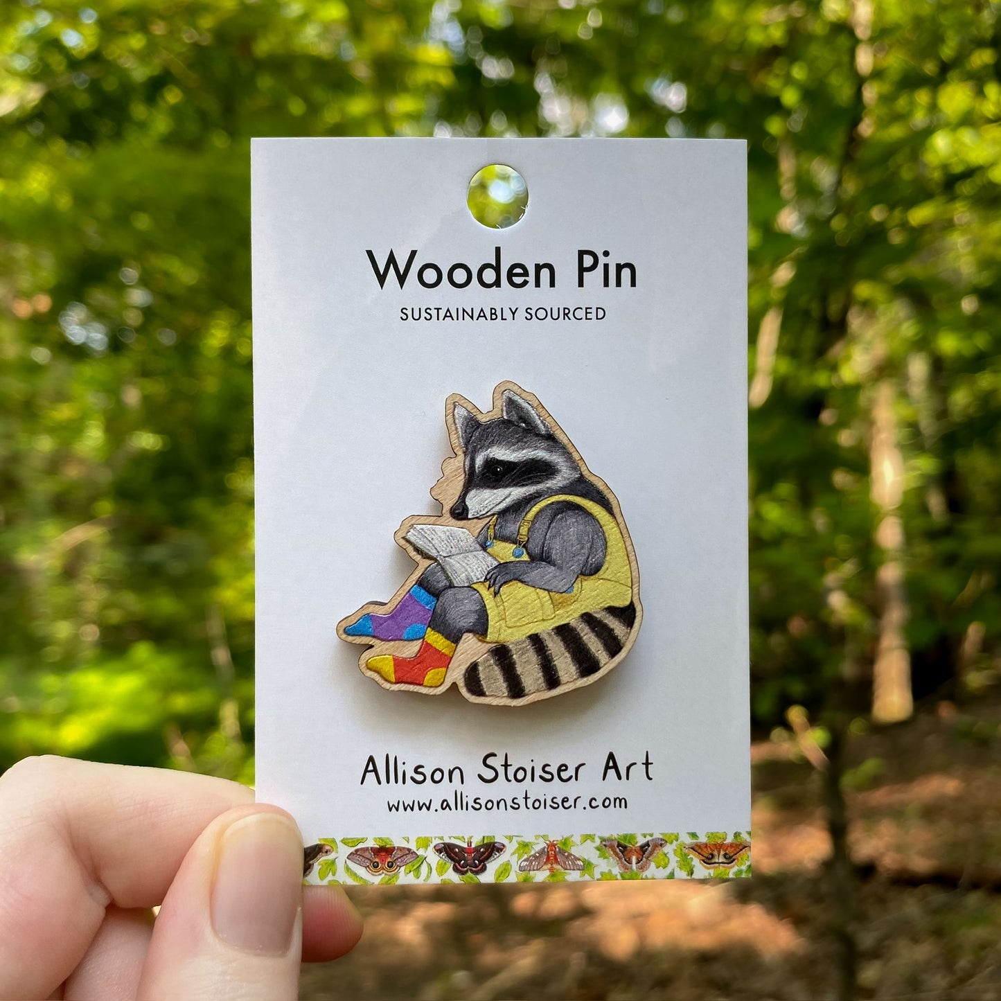 Wooden pin of a raccoon reading a book on a card with 'Allison Stoiser Art' branding, held in front of a forest background.