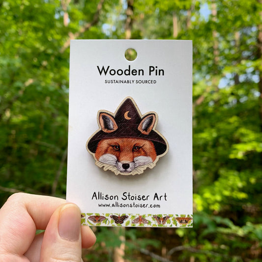 Wooden pin of a fox wearing a witch hat on a card with 'Allison Stoiser Art' branding, held against a forest background.