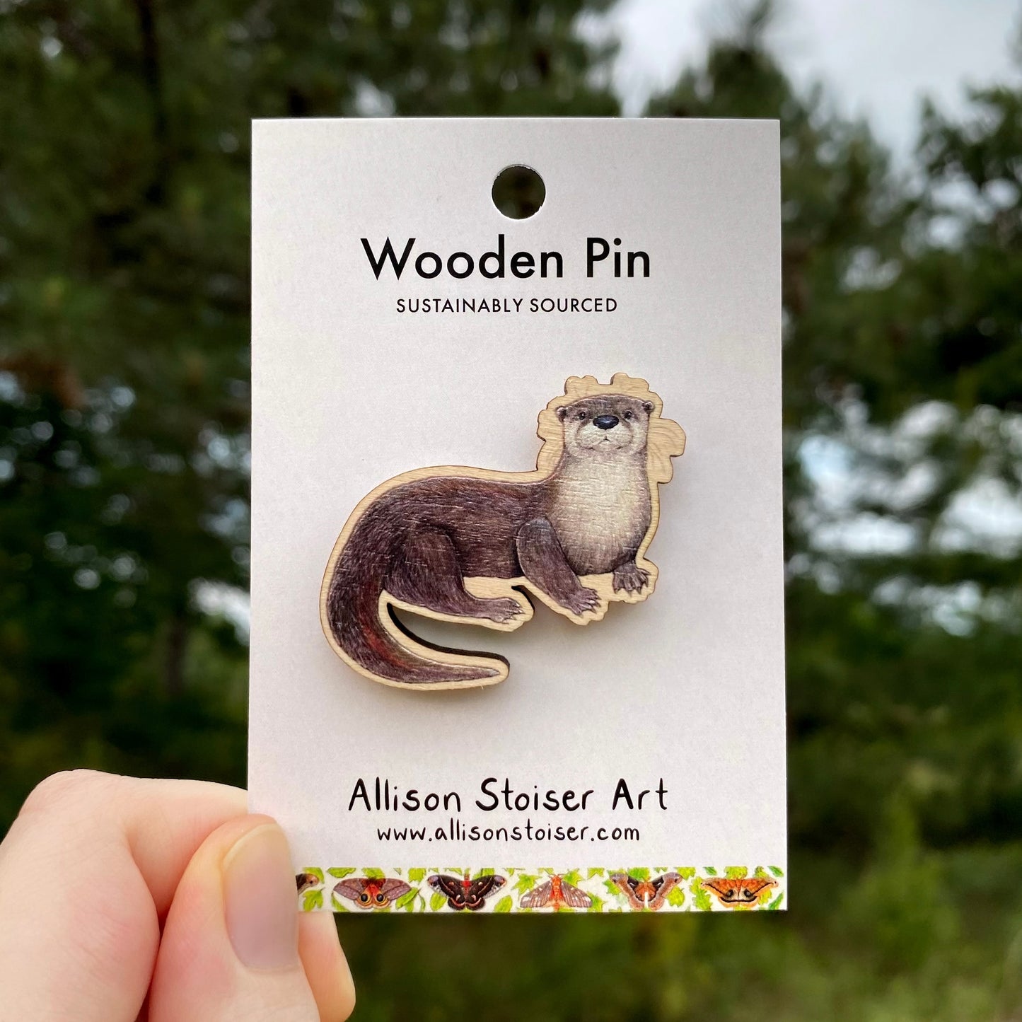 Wooden pin of an otter on a card with 'Allison Stoiser Art' branding, held against a blurred natural background.