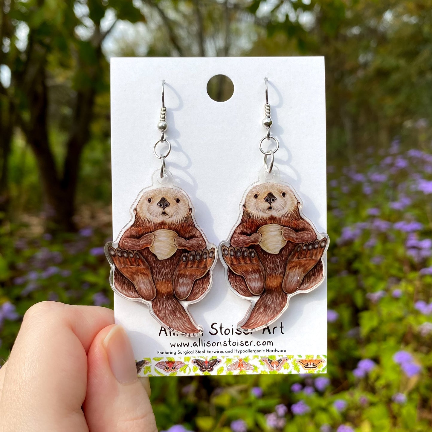 Pair of sea otter earrings on a card held in front of a natural background with flowers and trees.