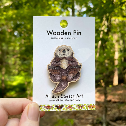 Wooden pin of a sea otter on a card with 'Allison Stoiser Art' branding, held against a forest background.