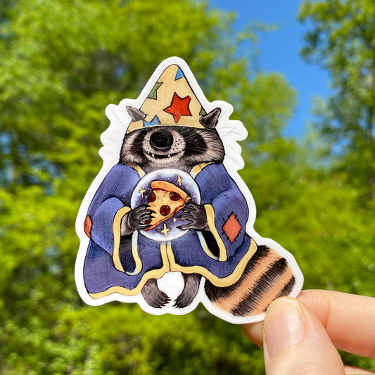 A vinyl sticker of a raccoon dressed as a sorcerer with a crystal ball foretelling of a pizza slice, held against a blurred background of trees.