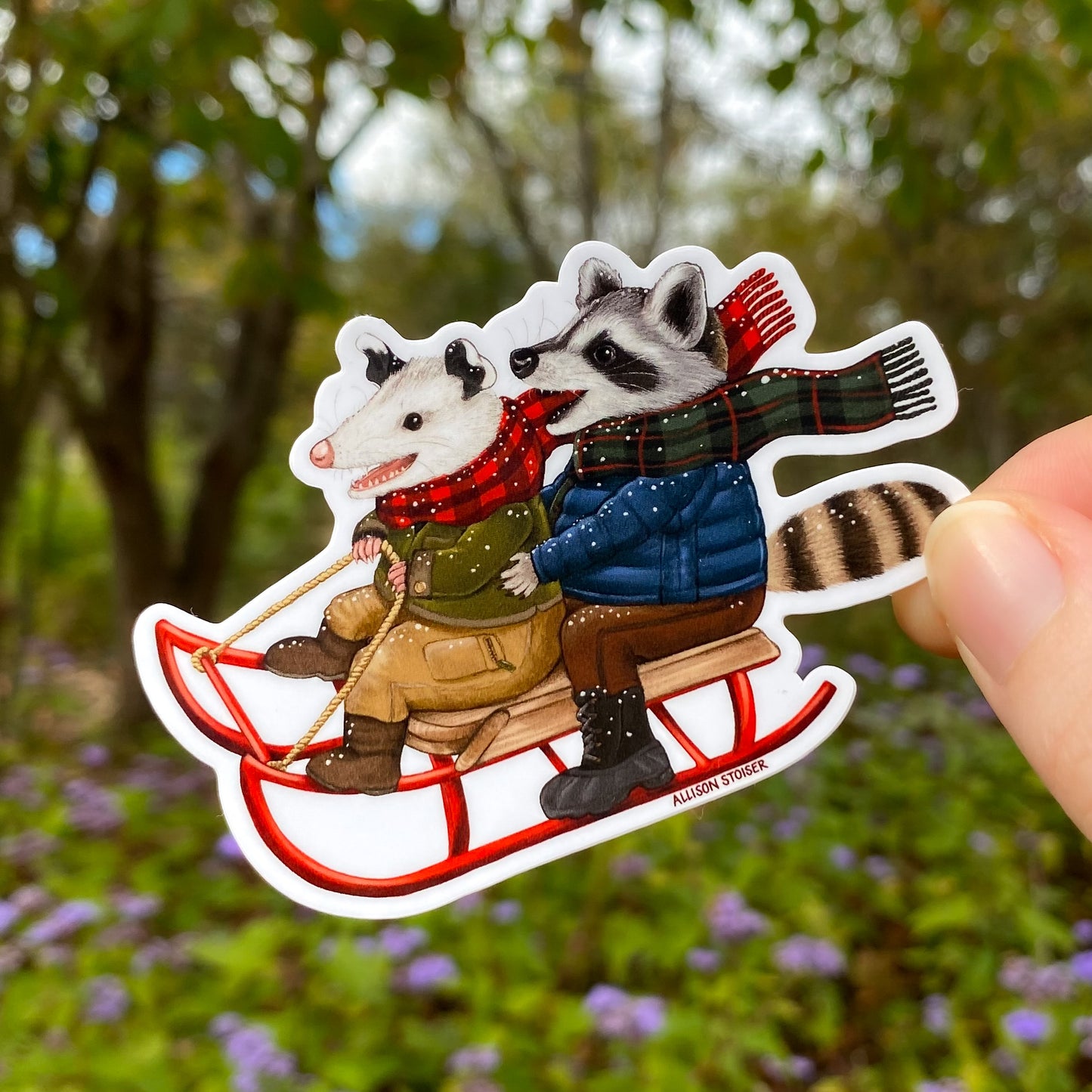 Sticker of an opossum and raccoon on a sled held in front of a natural background