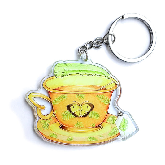 Double-sided acrylic keychain with an illustration of a sleepy orange caterpillar inside a yellow teacup, with green leaves and a butterfly on the saucer.