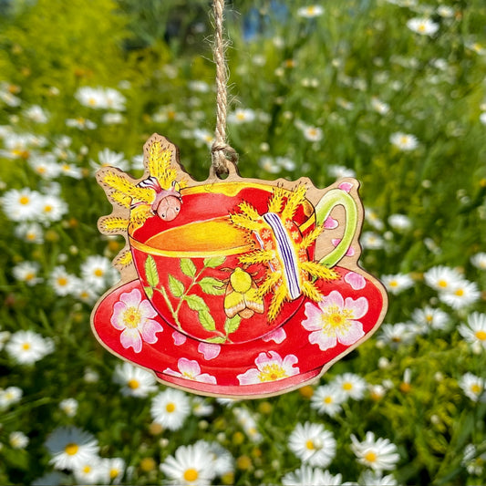 Decorative red teacup ornament with floral design and caterpillars sitting at the rim hanging against a natural background with flowers.