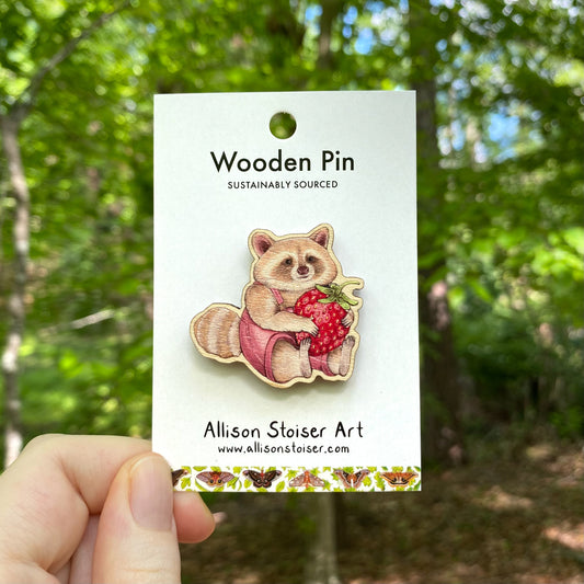 A wooden pin of a blond raccoon holding a strawberry
