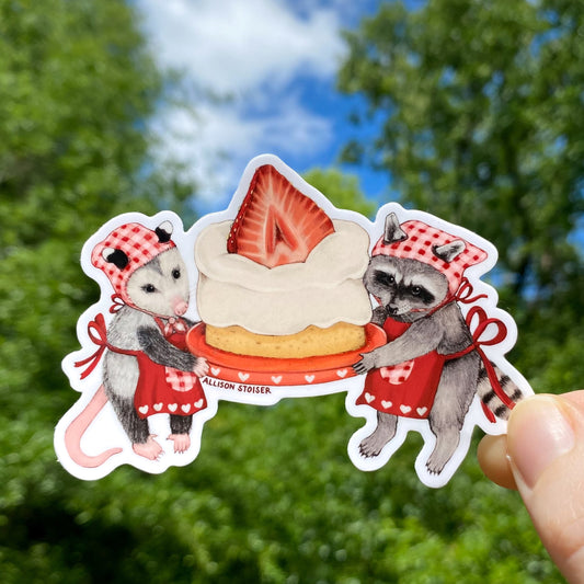 A hand holding a vinyl sticker of a raccoon and an opossum holding a cake