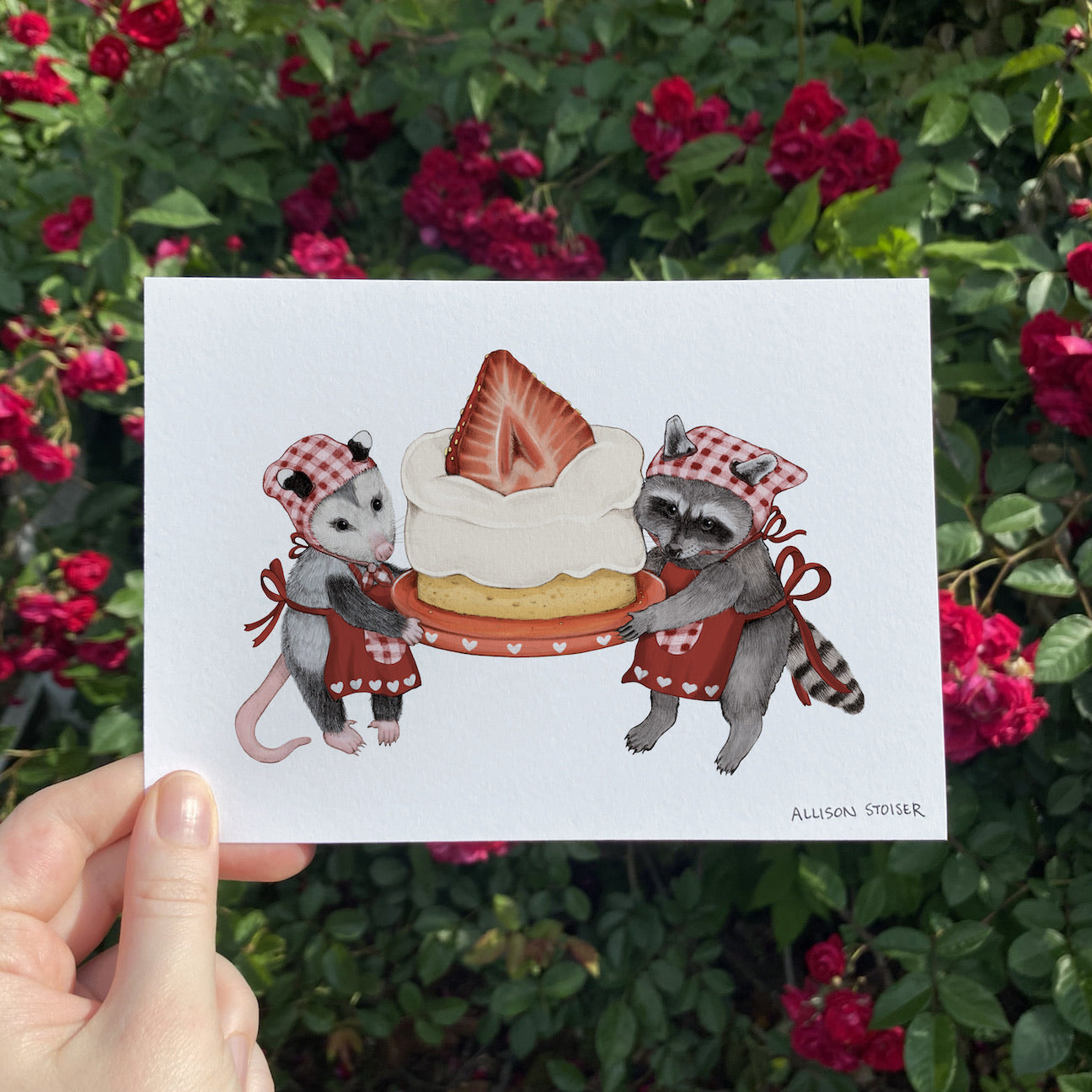 A hand holding a print of an opossum and raccoon holding a strawberry shortcake