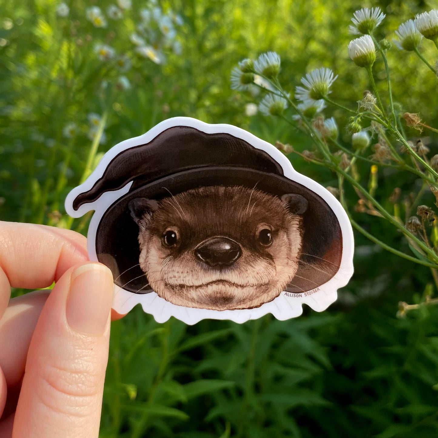 Vinyl sticker of an otter in a black wizard hat held in front of a background with flowers