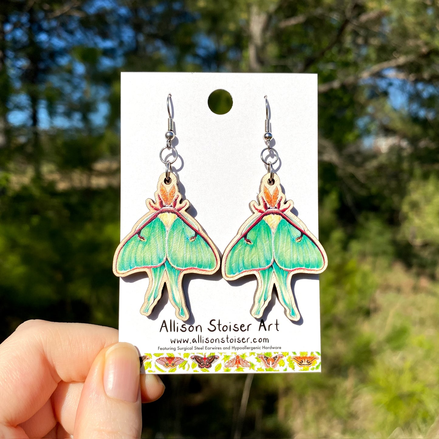 A pair of Luna moth earrings printed on birch plywood. The earrings are held by a hand against a blurred background of greenery.