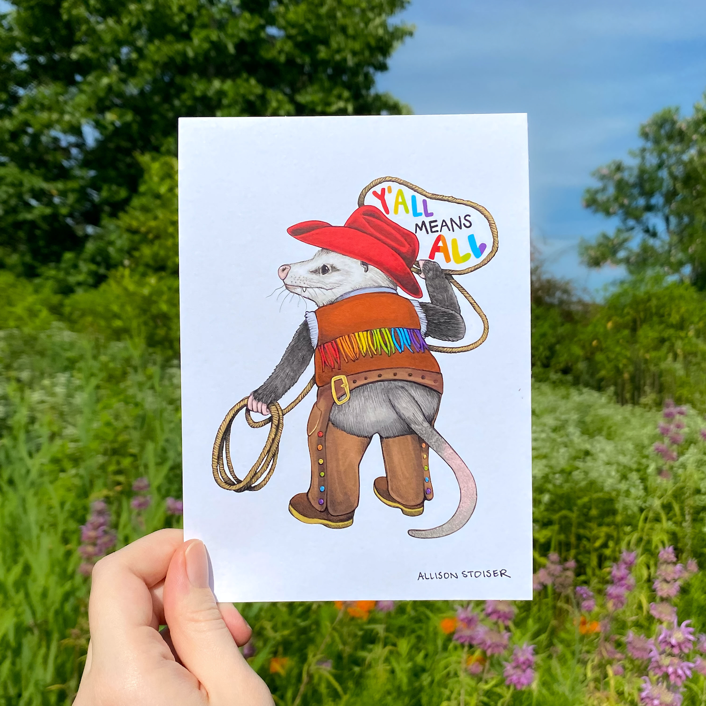 Illustration of an opossum in cowboy attire with 'Y'all Means All' text, held against a natural background.