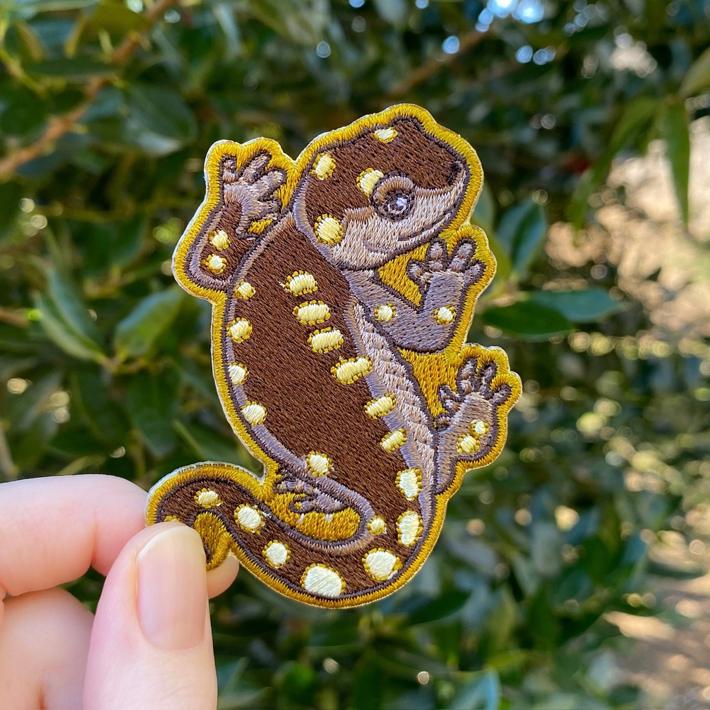 An embroidered patch of a yellow-spotted salamander held against a blurred background of greenery.