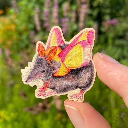 A sustainably sourced wooden pin featuring an illustration of a shrew dressed as a rosy maple moth