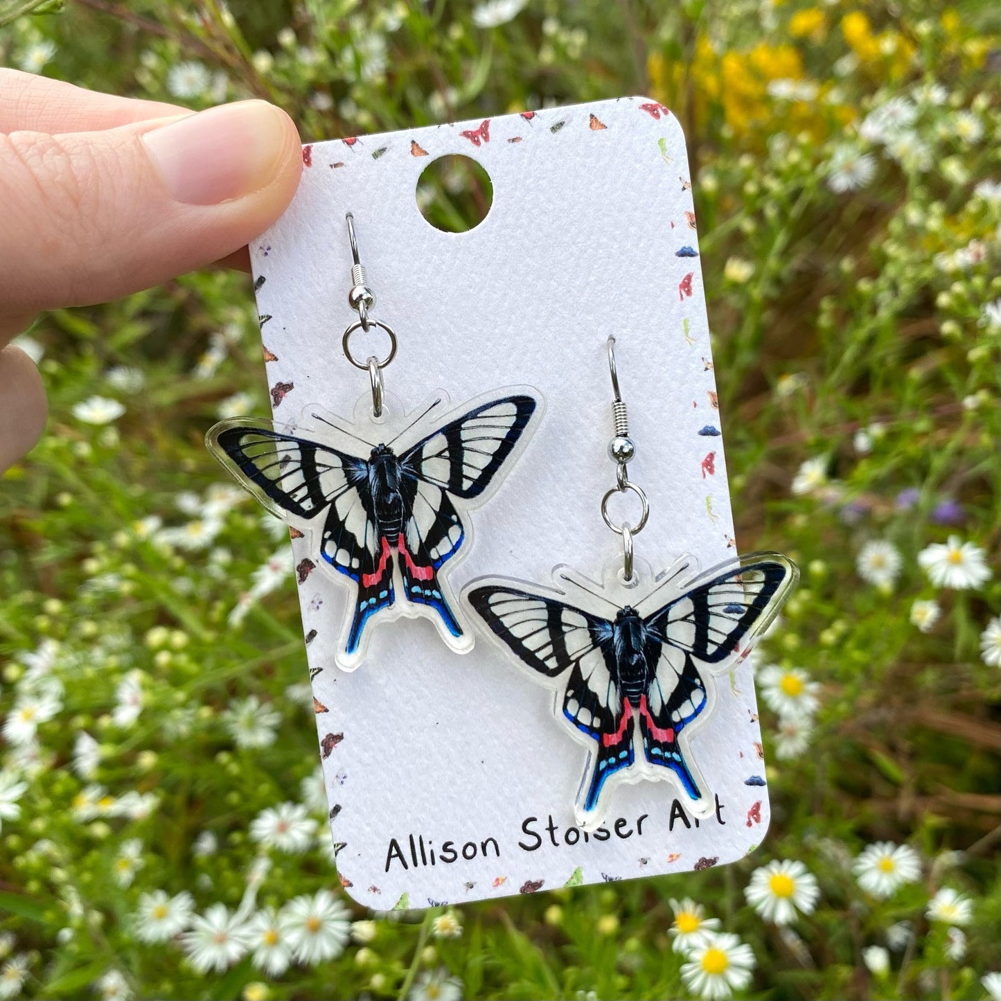 A hand holding a pair of acrylic earrings of sylphina metalmarks