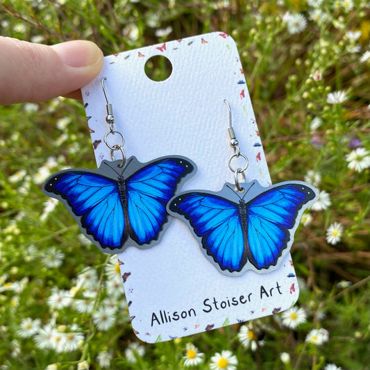 A hand holding a pair of metal morpho butterfly earrings