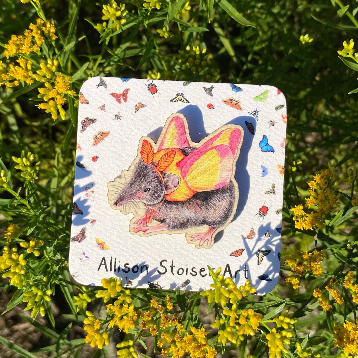 A sustainably sourced wooden pin featuring an illustration of a shrew dressed as a rosy maple moth