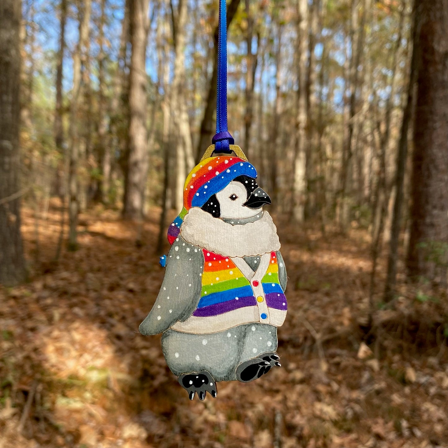 A hand-painted ornament of a penguin wearing a rainbow striped hat and sweater vest.