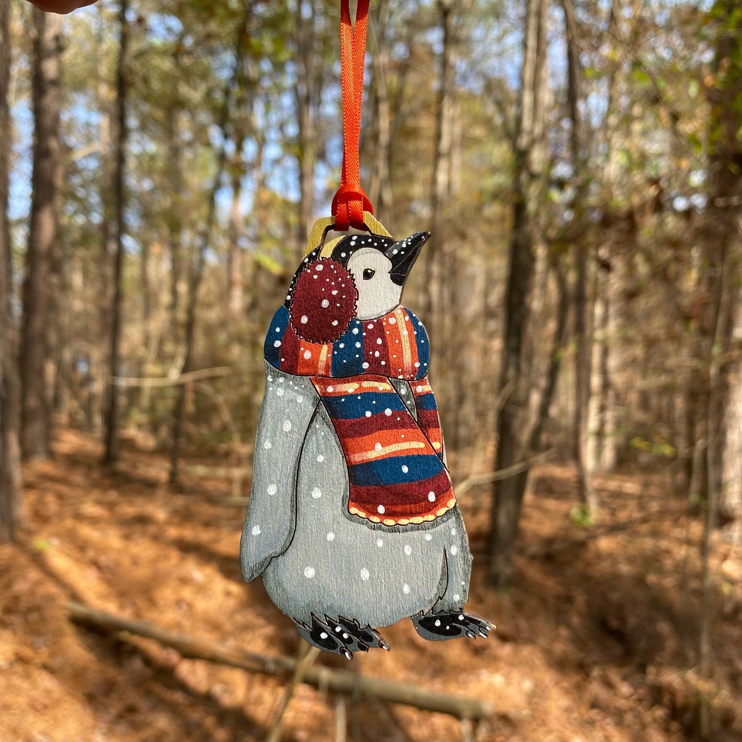 A hand-painted ornament of a penguin wearing a red earmuffs and a striped scarf