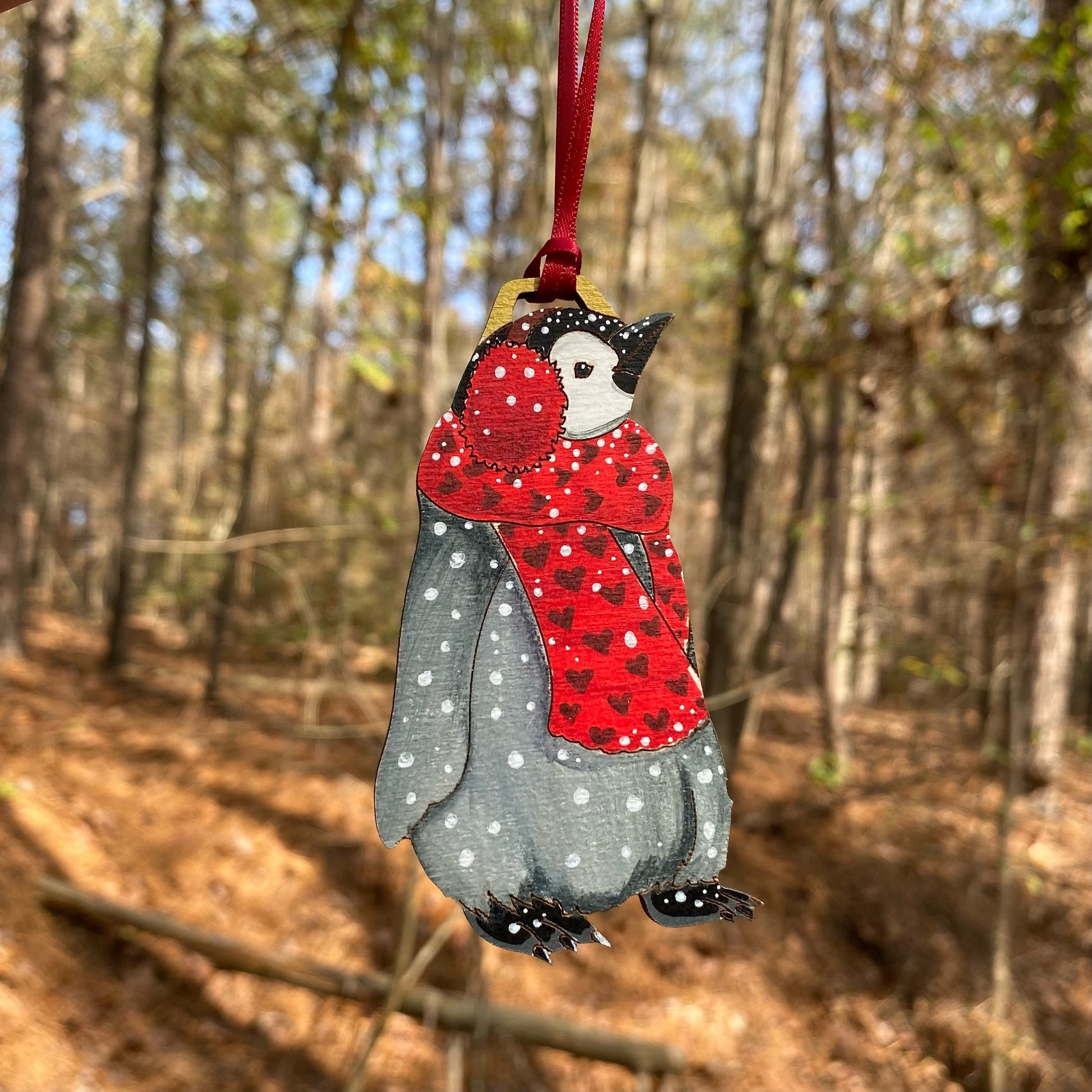 A hand-painted ornament of a penguin wearing a red earmuffs and a heart-print scarf