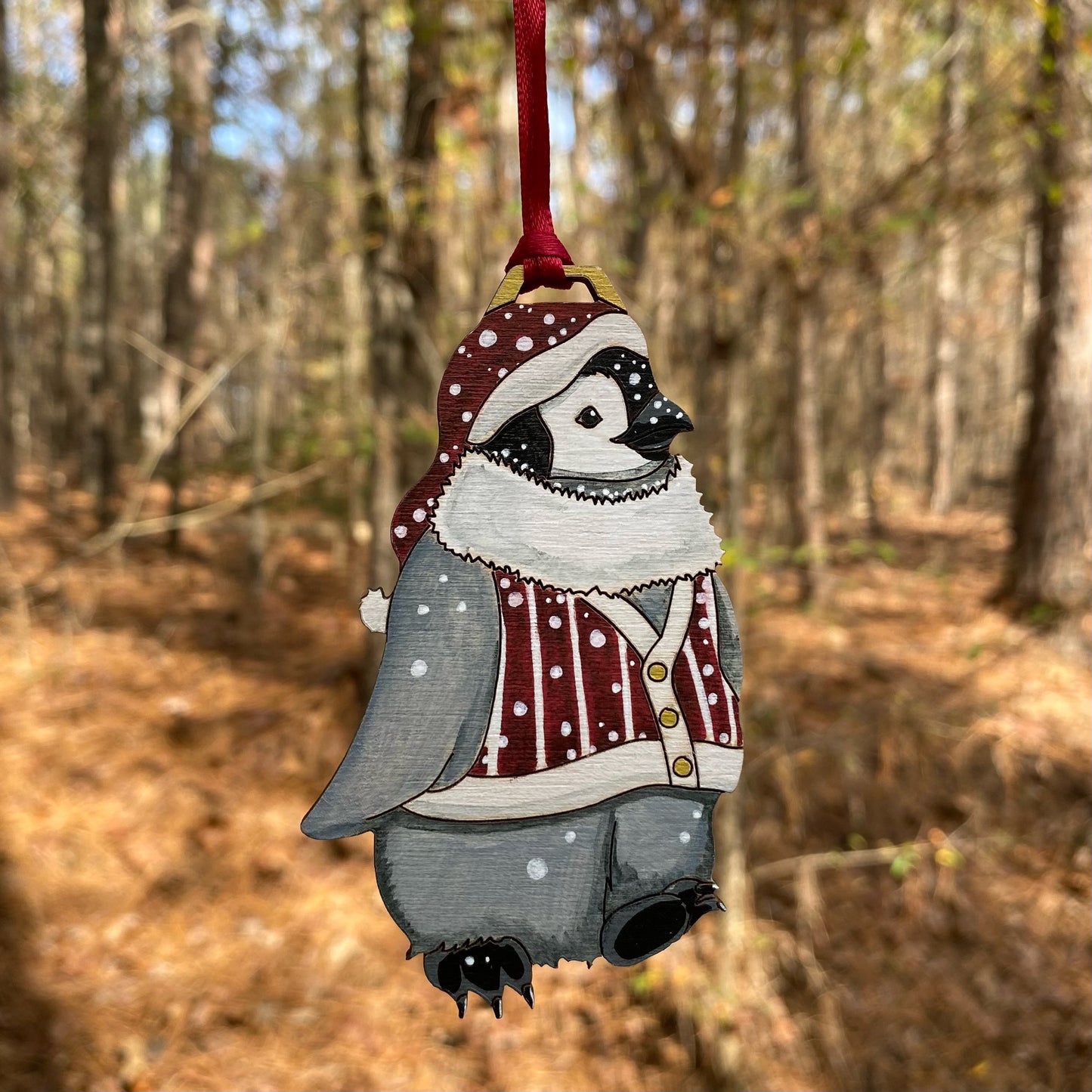 A hand-painted ornament of a penguin wearing a burgundy hat and sweater vest.