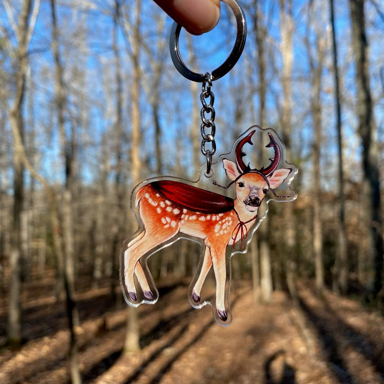 A keychain of a deer wearing stag beetle horns and a cloak being held in front of the forest.