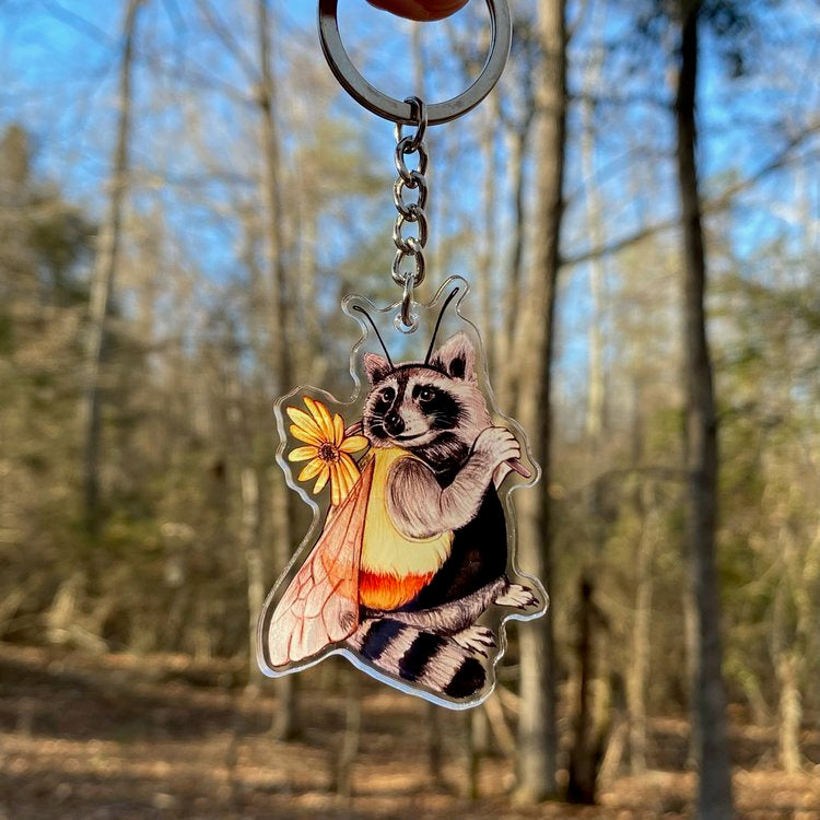A keychain of a raccoon wearing a bumble bee costume being held in front of the forest.