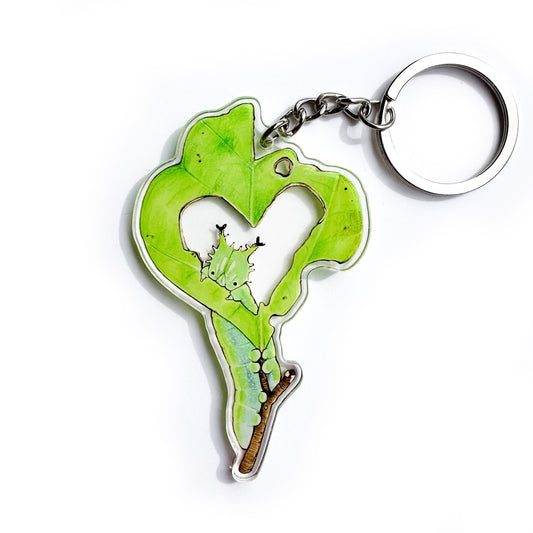 A keychain of a leaf with a heart-shaped hole and a caterpillar peeking out on a white background.