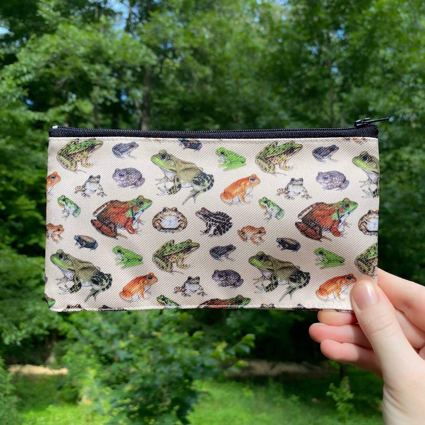 A hand holding a beige pouch with a pattern of frogs printed on it.