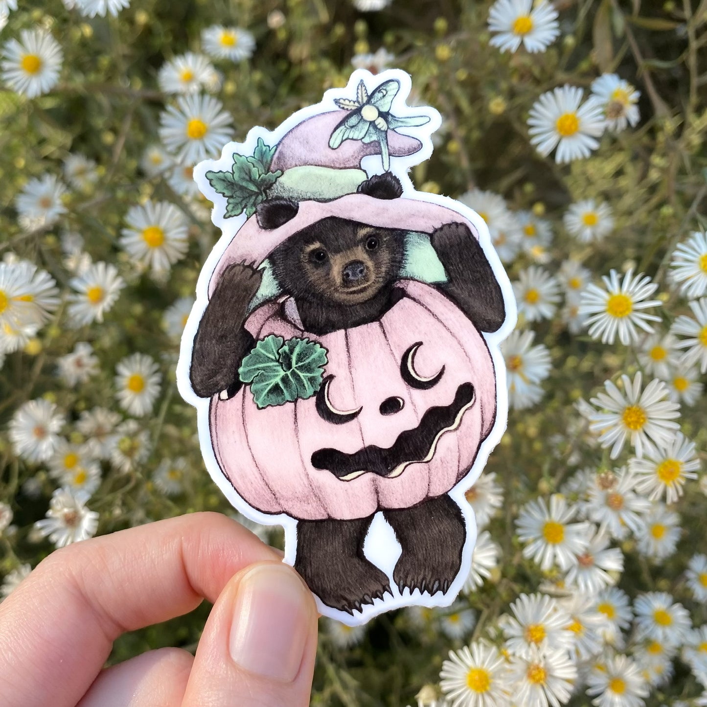 A hand holding a weatherproof vinyl sticker of a black bear cub in a pink pumpkin and witch hat.