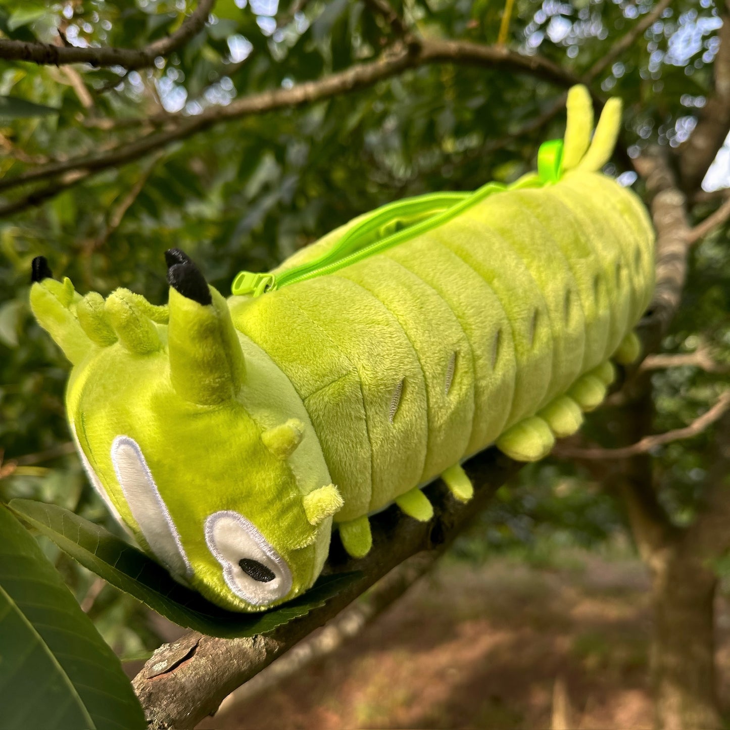 A plush caterpillar toy in bright lime green and yellow-green, resting on a leaf on a tree branch outdoors.