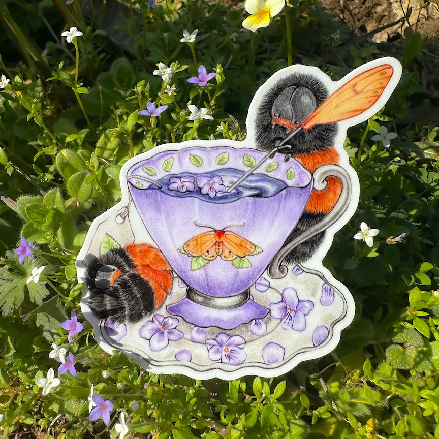 A hand holding a sticker featuring an illustration of a woolly bear caterpillar tea party