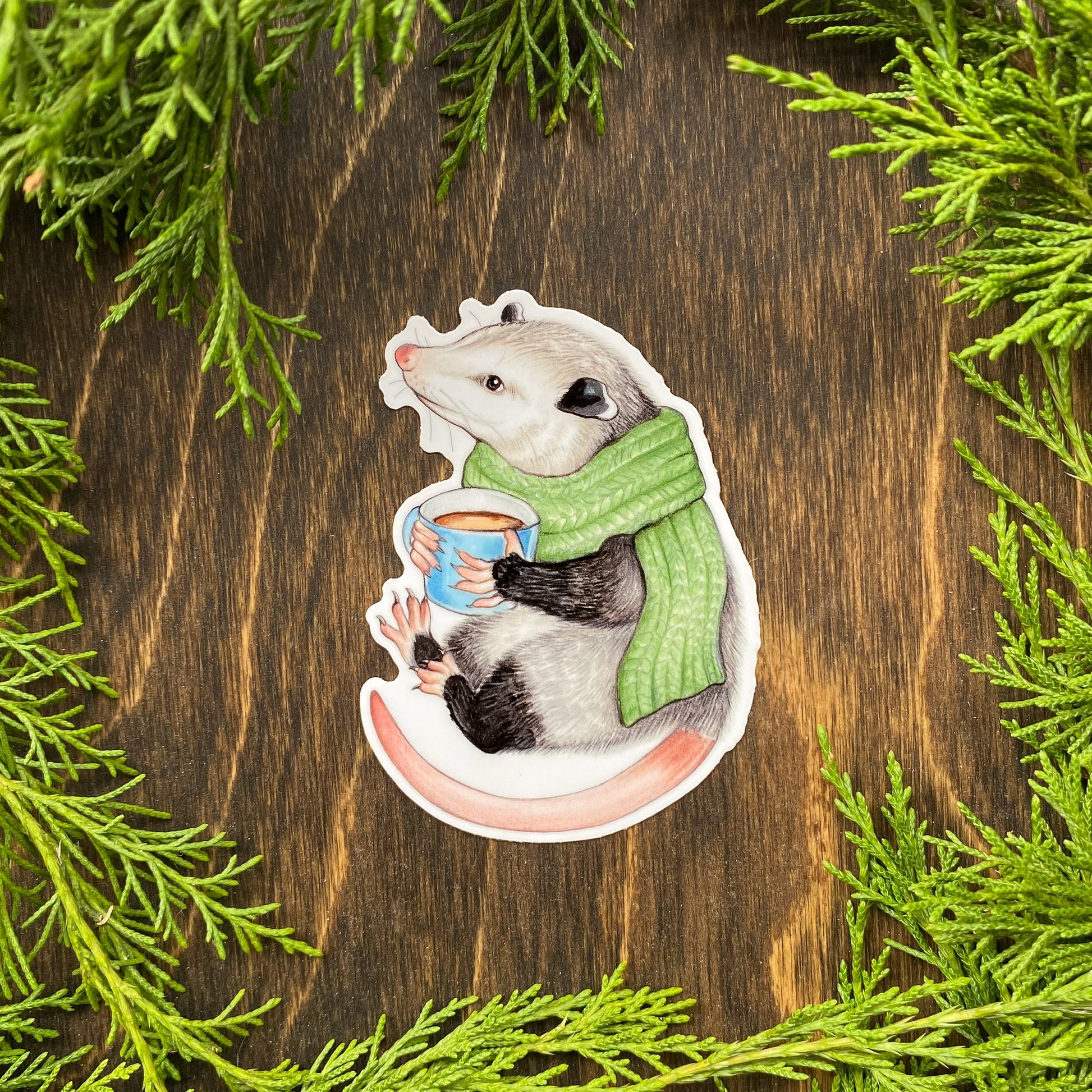 A weatherproof sticker featuring an illustration of a cozy opossum wearing a scarf and holding a mug of hot cocoa