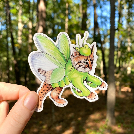 A hand holding a weatherproof vinyl sticker of a bobcat dressed up as a green praying mantis in front of a forest.