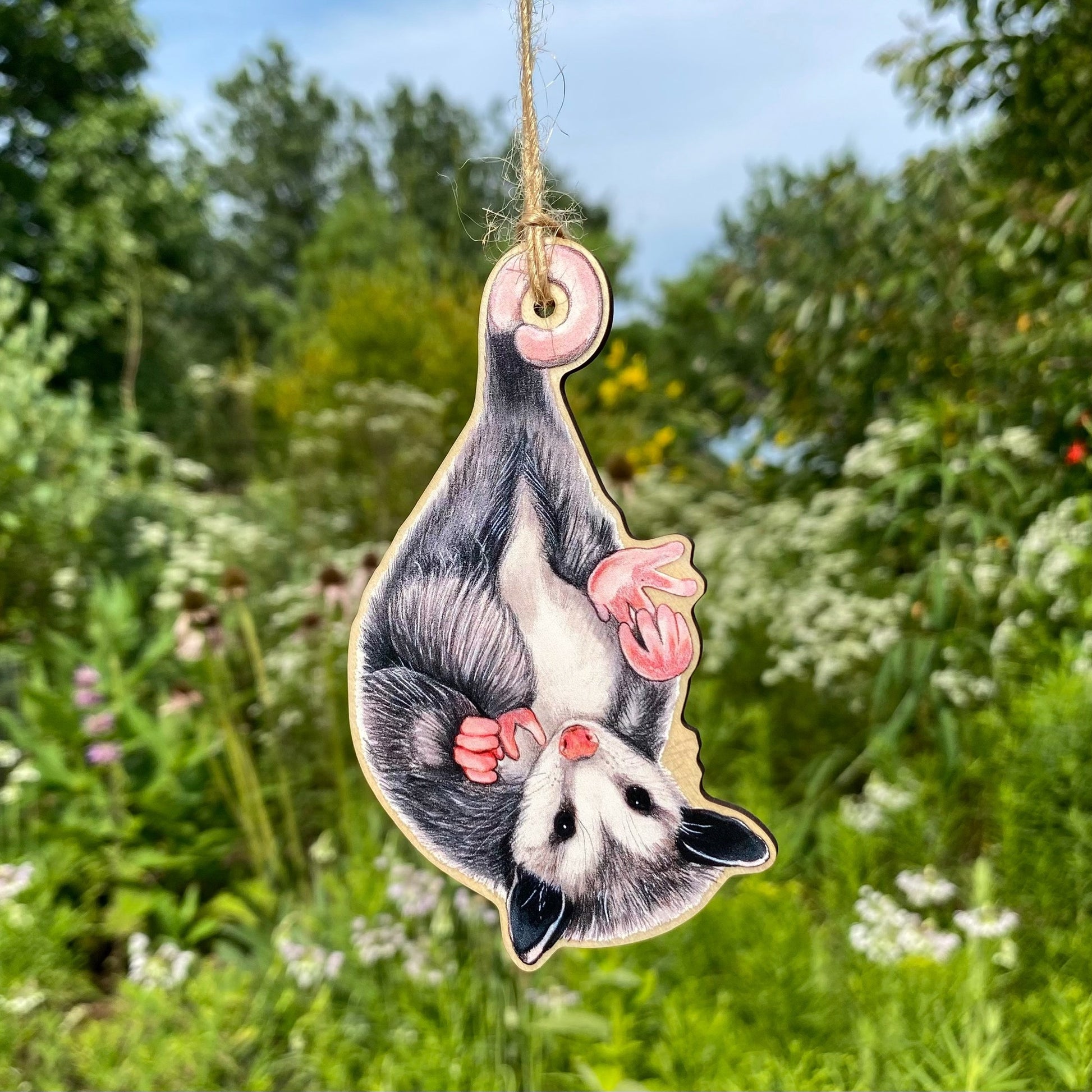 A wooden ornament strung with twine of a baby opossum hanging from its tail and holding onto one of its feet.