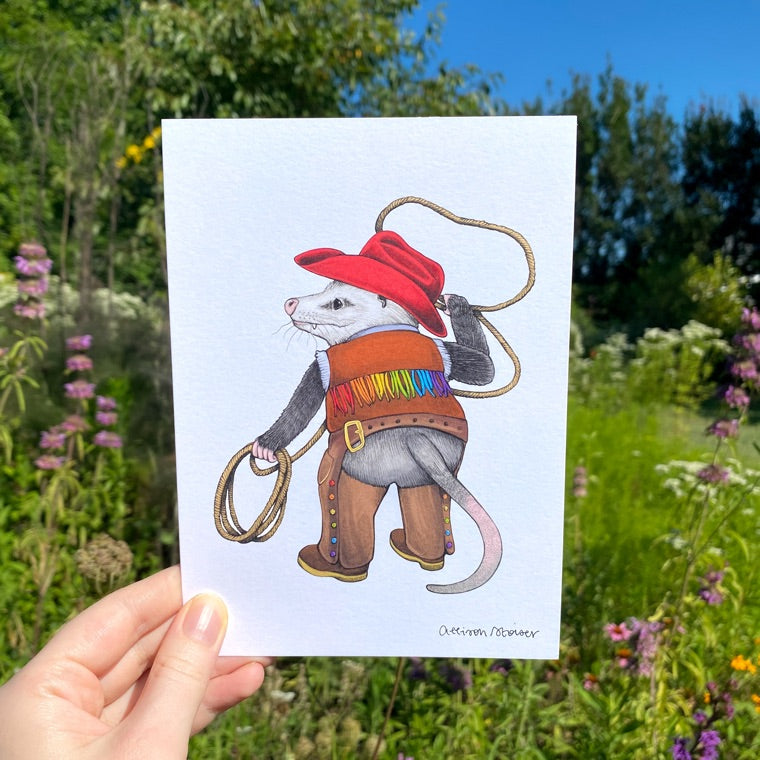 A hand holding a print of an opossum with a lasso dressed up in a rainbow cowboy outfit.