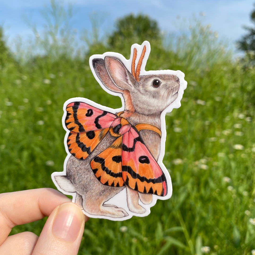 A hand holding a weatherproof vinyl sticker of a desert cottontail rabbit dressed up as a sheep moth.