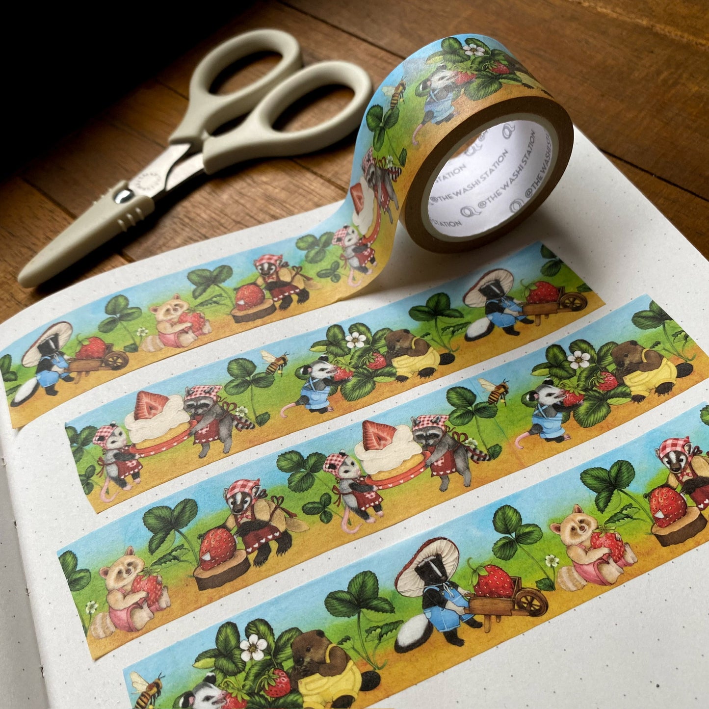 Washi tape of a farm scene with an opossum picking strawberries that a skunk takes in a wheelbarrow to a badger, who cuts the strawberry. Then there are a raccoon and opossum holding a cake. The washi tape measures 25mm and is swatched on a notebook.