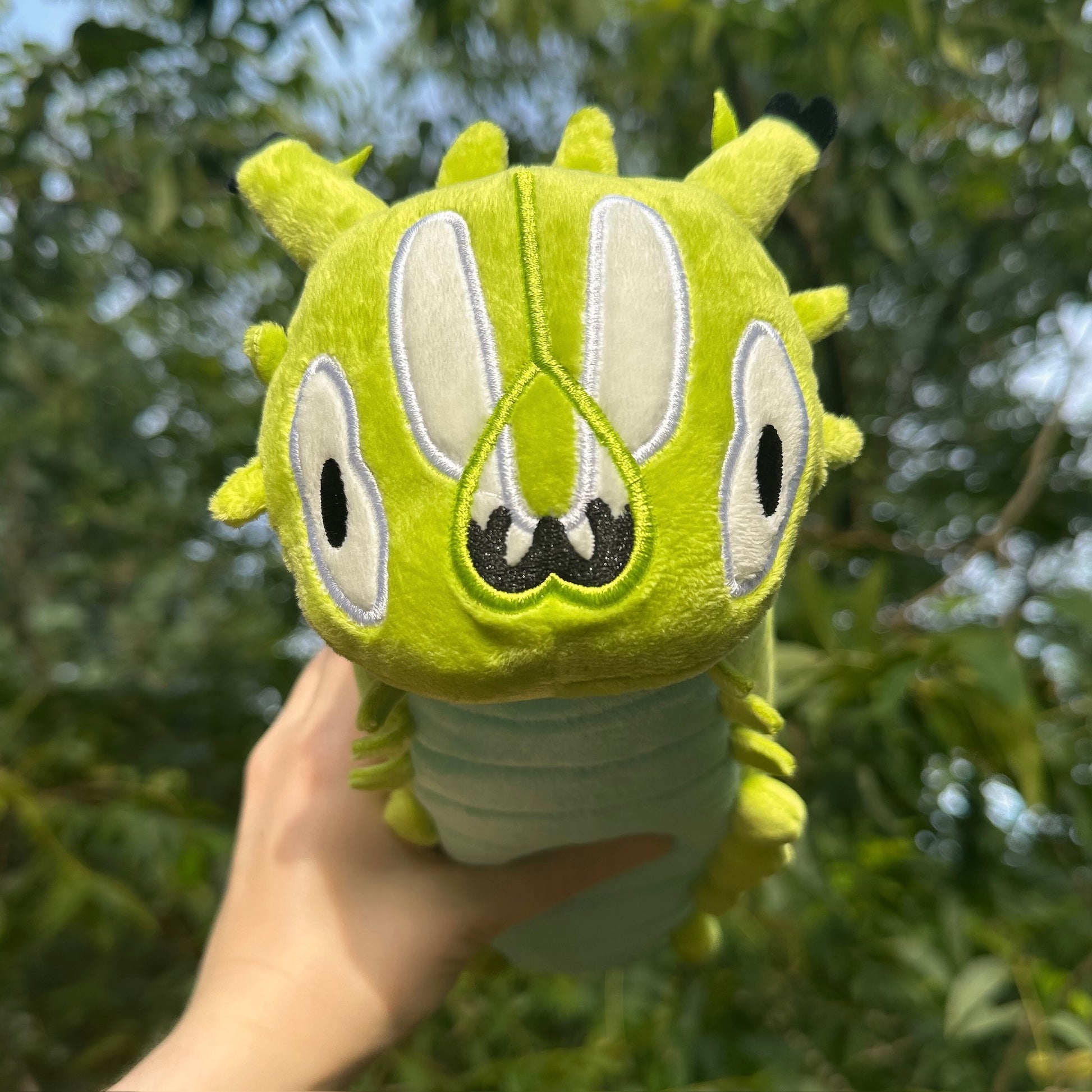 A green plush caterpillar pencil pouch being held up by a hand against a blurred outdoor background of green foliage.
