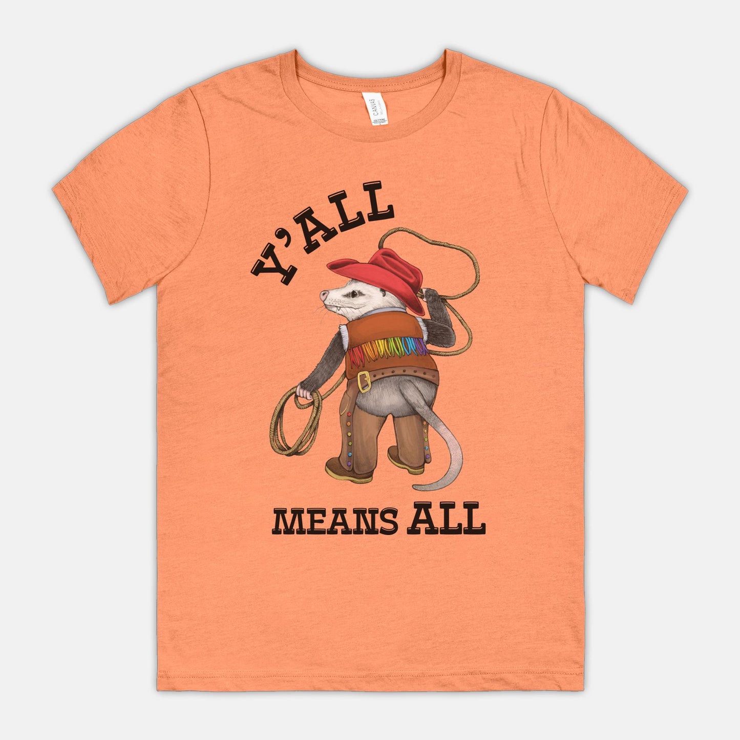 A peach-colored shirt featuring a cowboy opossum and text reading “Y’all Means All”