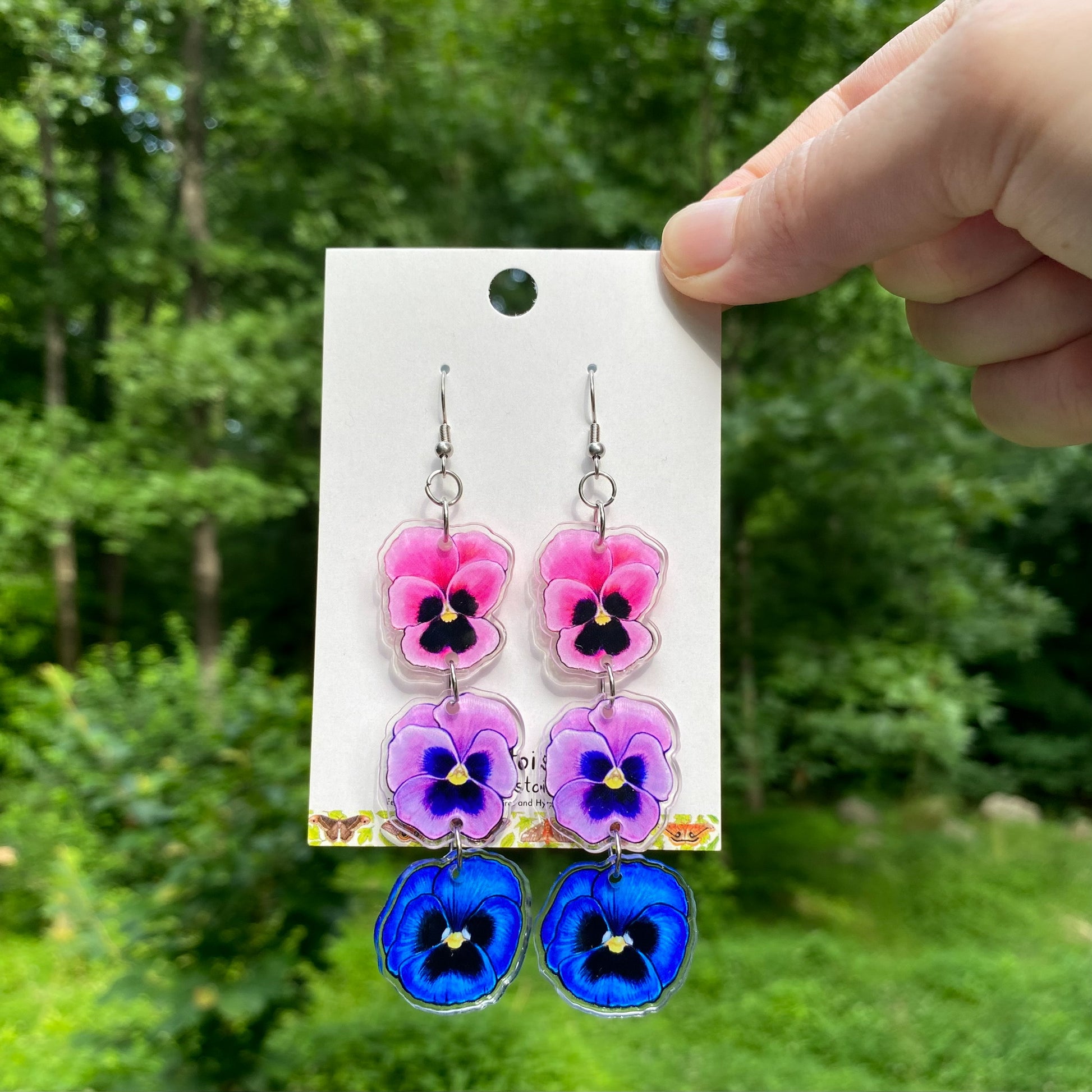 A hand holding a pair of acrylic earrings of pink, purple, and blue pansies.