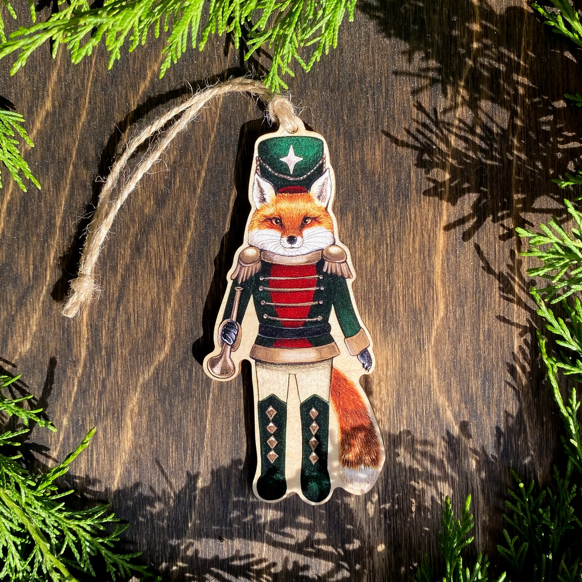 A wooden ornament strung with twine of a red fox in a red and green nutcracker outfit.