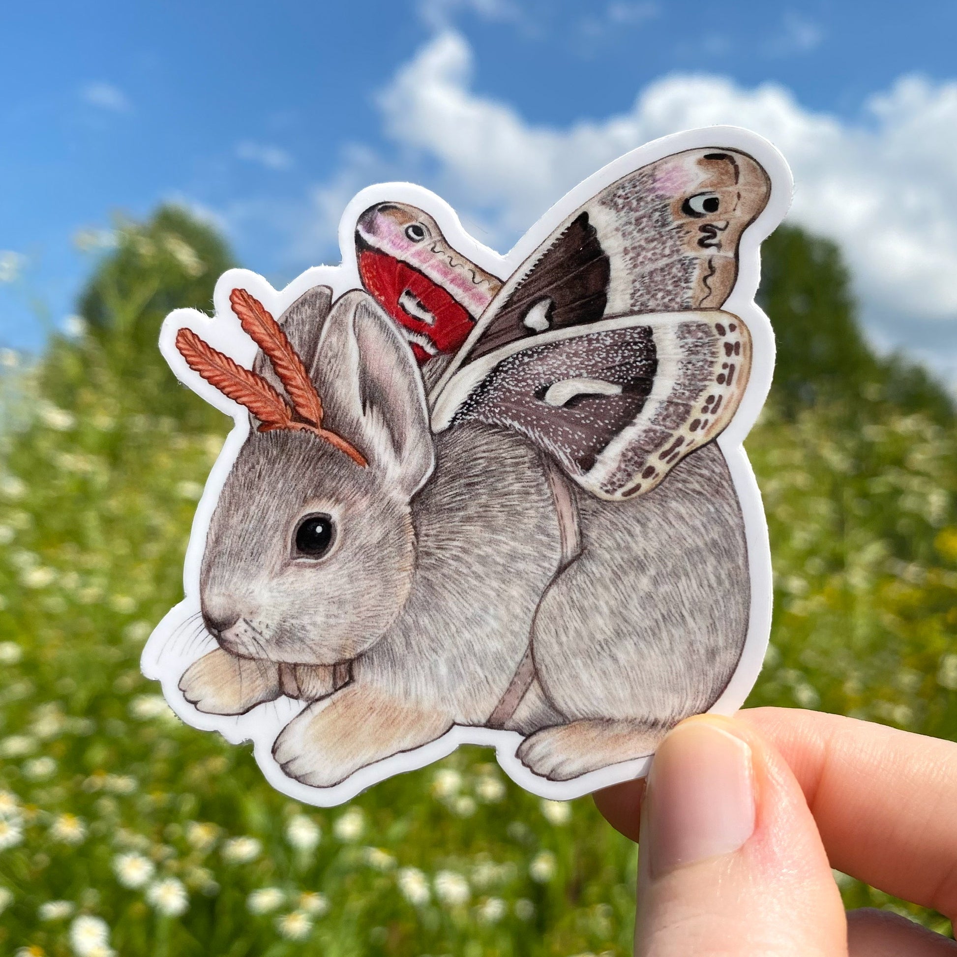 A hand holding a weatherproof vinyl sticker of a pygmy rabbit with Glover's silkmoth wings.