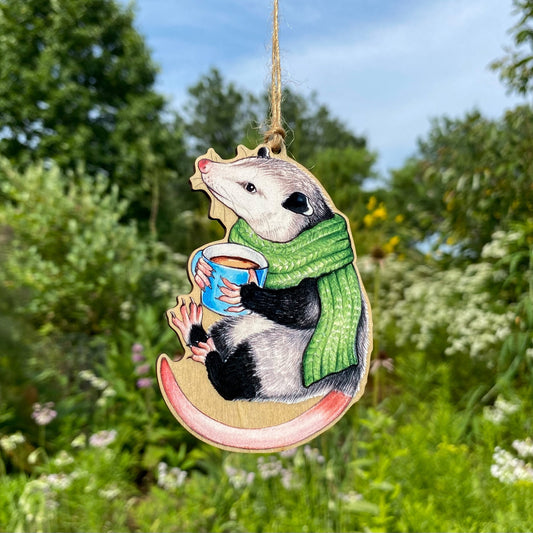 A wooden ornament strung with twine of an opossum in a green scarf and holding a mug of hot chocolate.