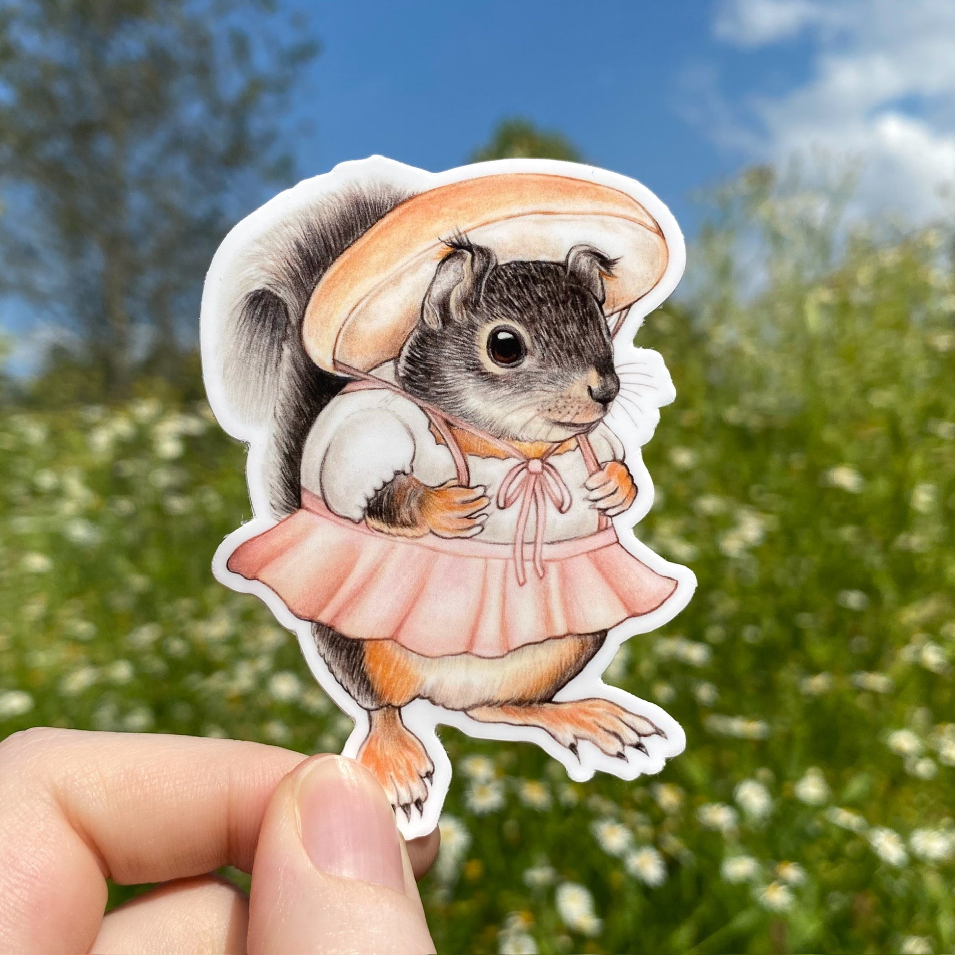 A hand holding a weatherproof vinyl sticker of a douglas squirrel with a porcini mushroom hat and skirt.