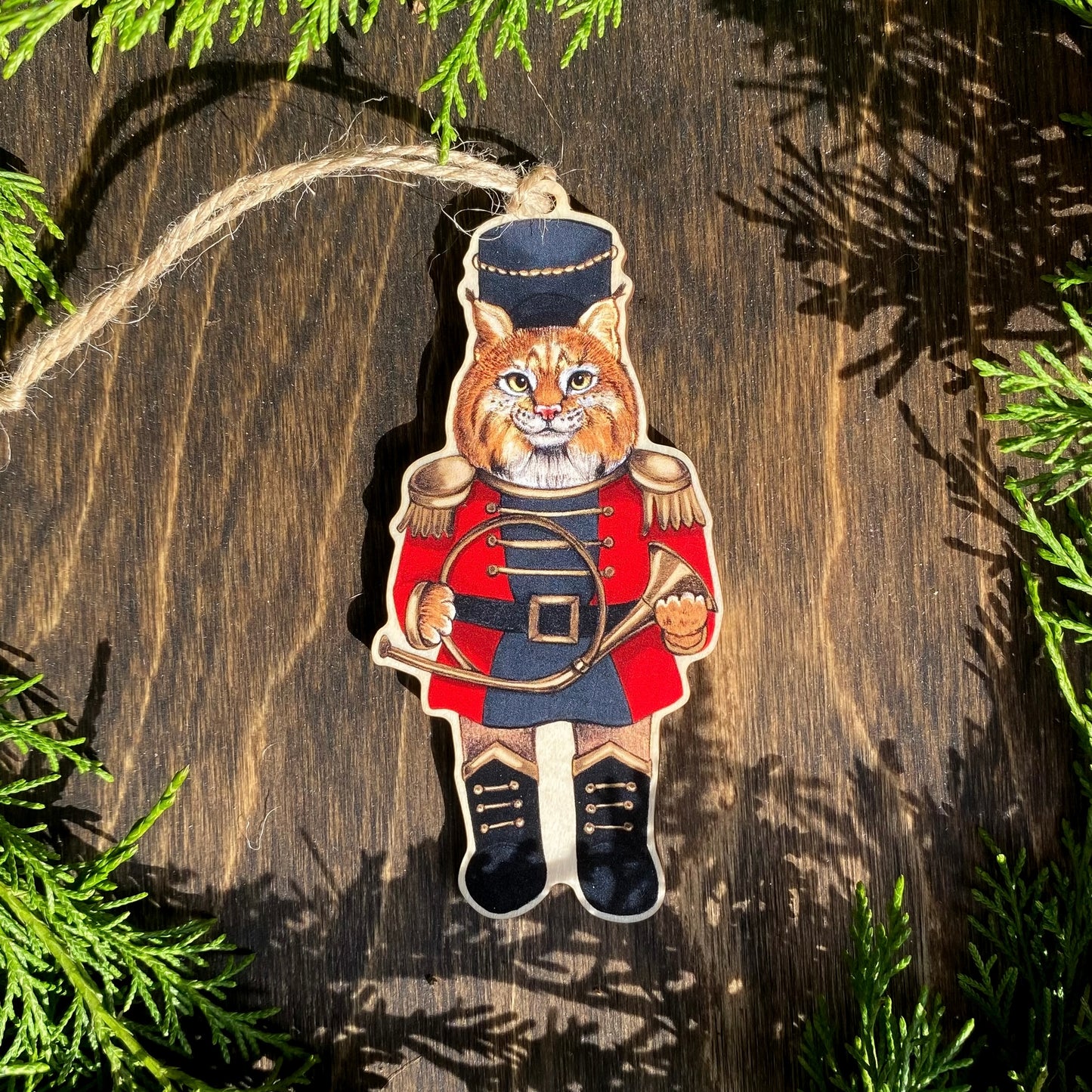 A wooden ornament strung with twine of a bobcat in a red nutcracker outfit.