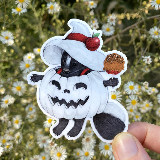 A hand holding a weatherproof vinyl sticker of a skunk in a white pumpkin and witch hat. The skunk is carrying a caramel apple.